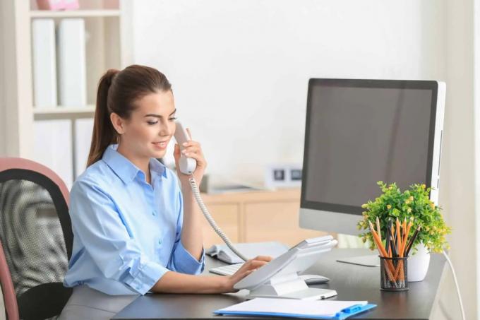 Moteris, sėdinti biure prie stalo, renka numerį telefonu 