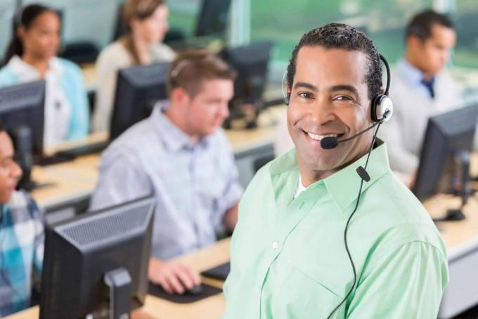 Muž s náhlavnou súpravou na úsmeve do kamery so zamestnancami call centra v pozadí